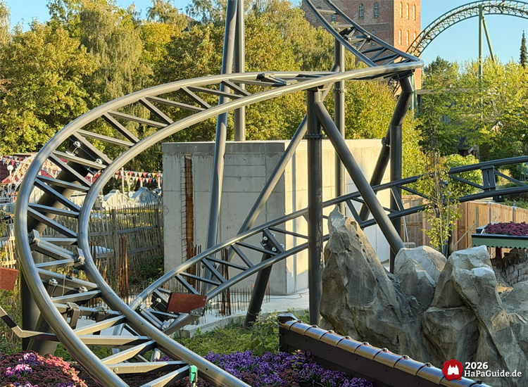 Stahlkonstruktion des Cornwall Coasters im Hansa-Park mit geschwungener Schiene und Betonwand des zukünftigen Roche Rocks, umgeben von Felsen und Blumen, im Hintergrund Bäume und weitere Streckenabschnitte
