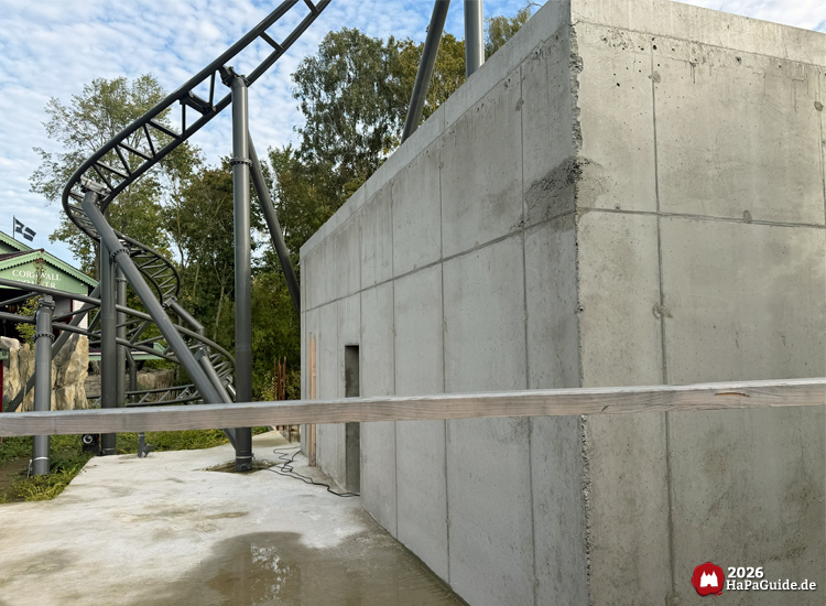 Großer Betonbau des zukünftigen Roche Rocks im Hansa-Park neben der Achterbahnstrecke des Cornwall Coasters, graue Wände mit sichtbaren Fugen, dahinter grüne Bäume und blauer Himmel mit leichten Wolken