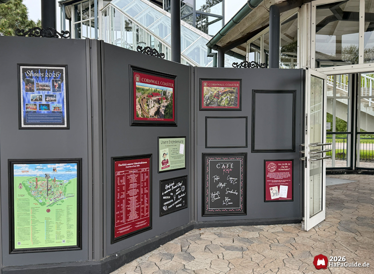 Informationswand auf dem Großparkplatz des Hansa-Park mit Plakaten zu Shows 2026, Cornwall Coaster, Parkplan, Café-Angeboten und Größenangaben vor dem verglasten Turmgebäude