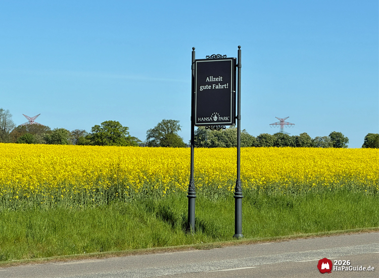 Schwarzes Hansa-Park Abschiedsschild mit der Aufschrift „Allzeit gute Fahrt!“ am Straßenrand vor einem leuchtend gelben Rapsfeld und blauem Himmel