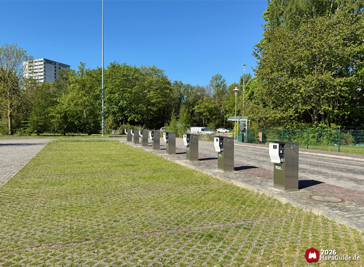 Reihe moderner Edelstahl-Ladesäulen auf dem E-Parkplatz des Hansa-Park, umgeben von grünen Bäumen und Rasenflächen bei klarem, sonnigem Himmel