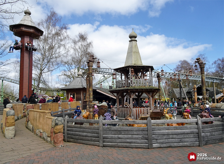 Peterhof von Novgorod im Hansa-Park mit Kinderkarussell Tierkindern vom Peterhof und dem Turm vom Peterhof, umgeben von rustikalen Gebäuden und Schienen der Flucht von Novgorod, Besuchende genießen die Attraktionen bei sonnigem Wetter
