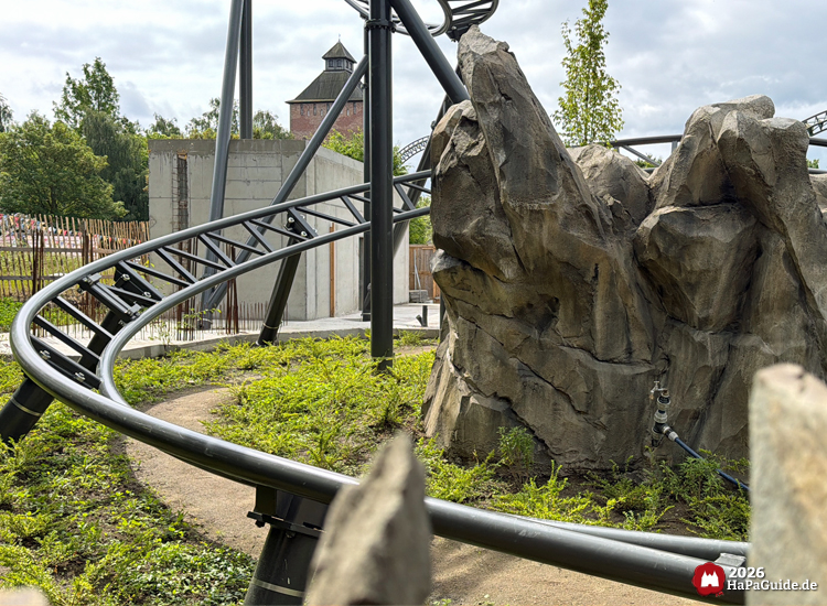 Baustelle der neuen Achterbahn Cornwall Coaster im Hansa-Park mit schwarzer Schienenkurve, künstlichen Felsen und grüner Bepflanzung, im Hintergrund der Saviour-Turm der Flucht von Novgorod