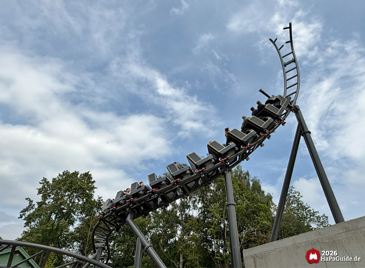 Zug des Cornwall Coasters im Hansa-Park fährt durch eine Kurve auf schwarzer Schiene den Spike hoch, getragen von Stahlstützen, vor grünem Baumbestand und blau-grauem Himmel