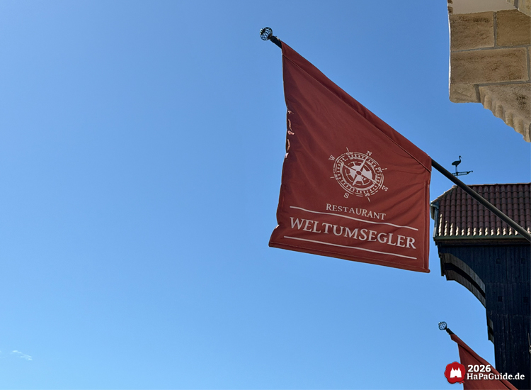 Rote Flagge mit der Aufschrift 'Restaurant Weltumsegler' und Kompasssymbol am Gebäude des Hansa-Park Restaurants, wehend vor klarem blauen Himmel