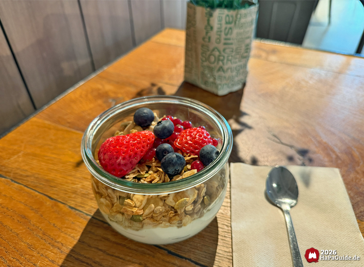 Glas mit Joghurt, Granola und frischen Beeren auf einem Holztisch im Restaurant Weltumsegler des Hansa-Parks, daneben ein Löffel und Serviette