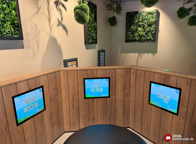 Kinderbereich im Restaurant Weltumsegler des Hansa-Parks mit drei interaktiven Bildschirmen und grüner Pflanzenwand, gestaltet mit Holzverkleidung und hängenden Mooskugeln