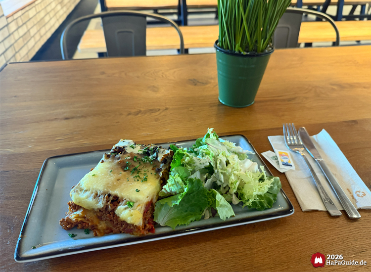 Tagesgericht Lasagne mit Käse und Kräutern, serviert mit grünem Salat auf grau-blauem Teller im Restaurant Weltumsegler des Hansa-Parks, auf einem Holztisch mit Besteck und Pflanzendekoration