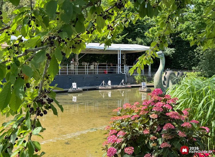 Teich der Fernlenkschiffe im Hansa-Park mit Bootsanleger und überdachtem Steg, im Vordergrund rosa Hortensien und grünes Laub, dahinter Figur des Seeungeheuers Nessie zwischen Pflanzen
