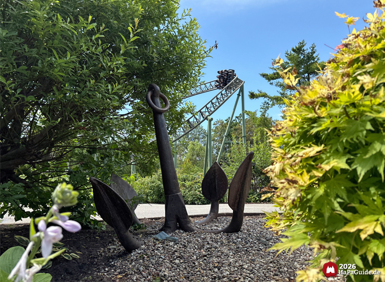 Großer Metallanker im Hansa-Garten des Hansa-Parks, umgeben von grünen Sträuchern und Kies, im Hintergrund Achterbahn Flucht von Novgorod mit Fahrgästen unter blauem Himmel
