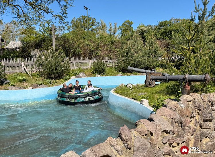 Boot der Wasserattraktion Störtebeker’s Kaperfahrt im Hansa-Park mit fünf Fahrgästen in einem grünen Rundboot, fährt durch einen blauen Wasserkanal mit Felsen und Kanone im Hintergrund