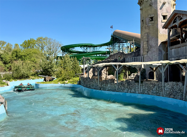 Wasserattraktion Störtebeker’s Kaperfahrt im Hansa-Park mit Rundboot in blauem Wasserkanal vor einer Steinwand mit Turm und Holzveranda, im Hintergrund der grüne Rutschkanal