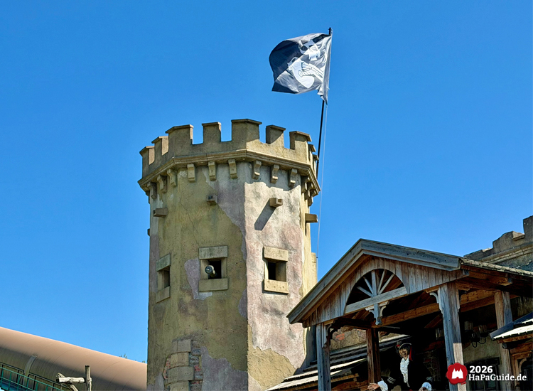 Turm der Wasserattraktion Störtebeker’s Kaperfahrt im Hansa-Park mit verwitterter Fassade und wehender Piratenflagge, daneben Holzveranda mit Piratenfigur Klaus Störtebeker unter klarem Himmel