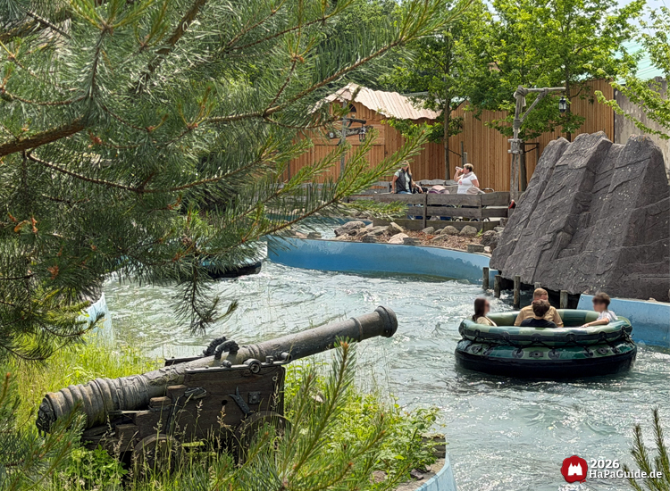 Piratenkanone aus Holz und Metall neben der Wasserbahn Störtebeker’s Kaperfahrt im Hansa-Park, Rundboot mit Fahrgästen fährt vorbei zwischen Felsen und Bäumen